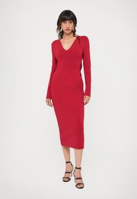 Punane kudum dresses V-kaelusega, pikkade varrukatega ja kehaümber oleva siluetiga, ulatudes keskmiste säärteni. Ühendatud mustade kõrgete kontsadega sandaalidega.