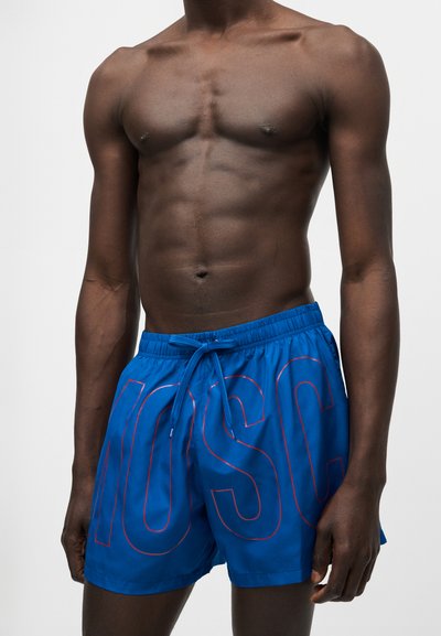 Άνδρας χωρίς πουκάμισο που φοράει μπλε μαγιό σορτς με μεγάλα κόκκινα γράμματα με περίγραμμα και κορδόνι στο μπροστινό μέρος, πάνω σε απλό φωτεινό φόντο.