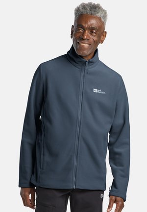 Lächelnder Mann, der eine dunkelblaue Jack Wolfskin Fleecejacke mit Reißverschluss und schwarze Hose trägt, vor einem einfarbigen hellen Hintergrund stehend.