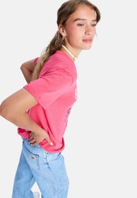 Ung kvinde i pink t-shirt og lyseblå jeans, med hænderne på lænden og kigger sidelæns mod en ensfarvet hvid baggrund.
