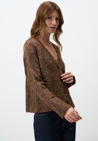Cardigan en tissu texturé marron avec un profond col en V, doté de trois boutons, de manches longues et d'une coupe décontractée. Porté sur un jean noir.
