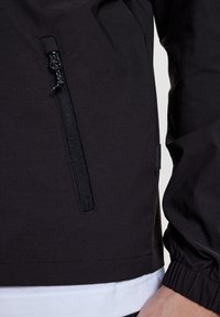 Černá bunda s kapsou na zip, elastickými manžetami, hladkou texturou a vrstevnatou bílou košilí pod ní.