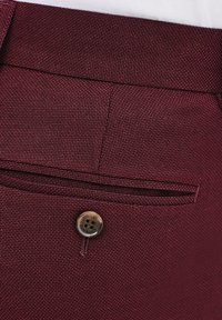 Burgundowe spodnie z teksturowanej tkaniny, z małą przednią kieszenią i okrągłym, metalowym guzikiem z wygrawerowanymi detalami.