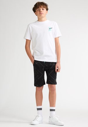 Maglietta bianca di cotone con logo turchese, pantaloncini neri con motivo geometrico e accenti verde neon, abbinati a sneakers bianche e calzini a righe.