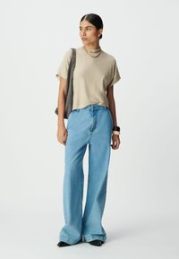 Beżowy, teksturowany t-shirt z krótkim rękawem w wzór chevron, zestawiony z jasnoniebieskimi dżinsami o szerokich nogawkach i czarnymi, spiczastymi butami. Szara torebka na ramię i biżuteria.