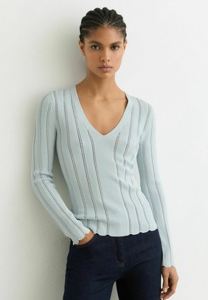 Nő, aki világoskék bordázott, V-nyakú pulóvert és sötét farmert visel, fonott hajjal, fehér fal mellett áll, kezét a zsebében tartva.