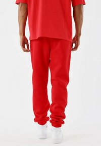 Man in rode trainingsbroek, rode T-shirt en witte sneakers, gezien vanaf de achterkant tegen een plain witte achtergrond.