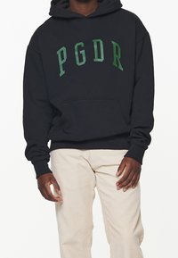 Felpa nera con cappuccio e lettering verde "PGDR", tasca frontale e polsini a coste, abbinata a pantaloni di corduroy beige chiaro.