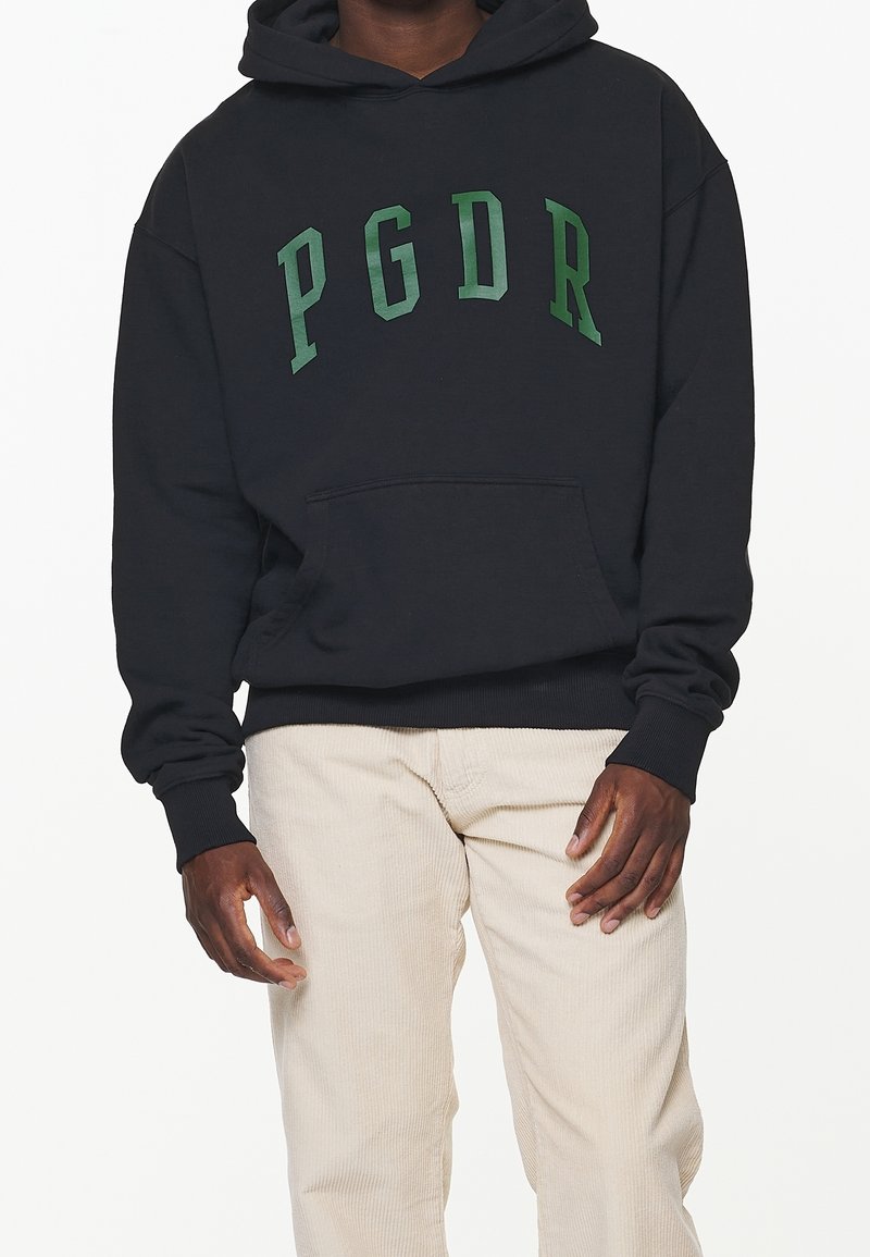 Felpa nera con cappuccio e lettering verde "PGDR", tasca frontale e polsini a coste, abbinata a pantaloni di corduroy beige chiaro.
