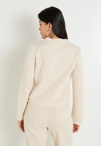 Top de manga larga beige con cuello redondo y textura acanalada, combinado con pantalones a juego que cuentan con bolsillos traseros. Diseño minimalista.