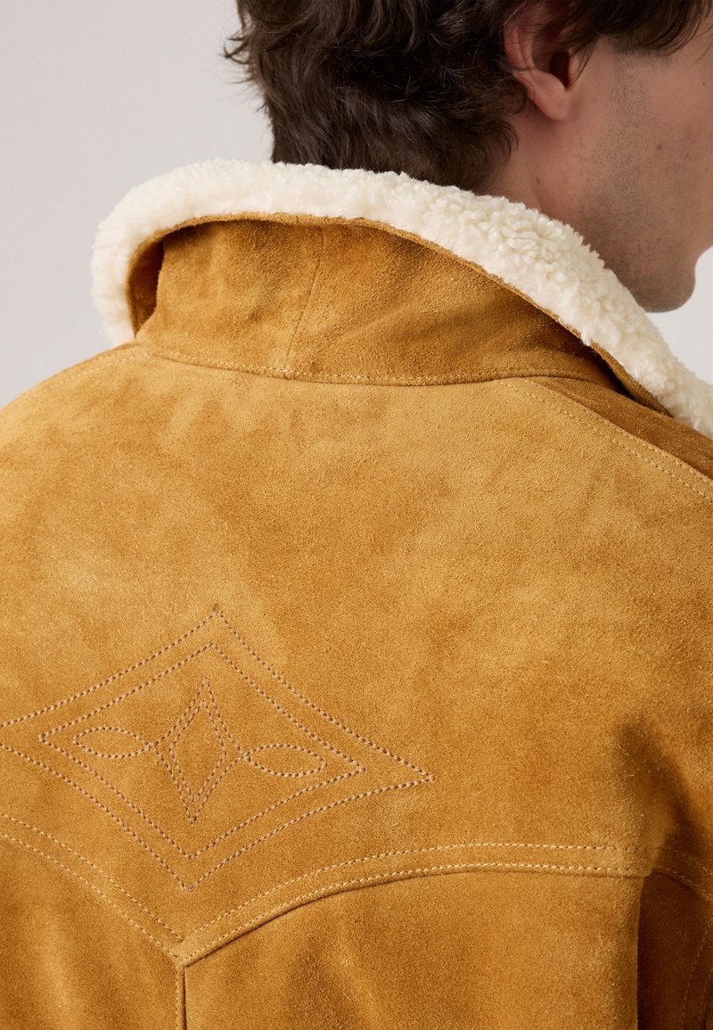 Bruine suède jas met een witte schapenvacht kraag. Bevat een ruitvormig gestikt detail op de achterkant. Zachte textuur en klassiek ontwerp.