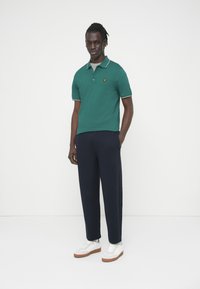 Πράσινο πόλο μπλουζάκι με αντίθετο γιακά, κεντημένο λογότυπο, μανίκια μέχρι τον αγκώνα· συνδυασμένο με σκούρο μπλε παντελόνι και λευκά αθλητικά παπούτσια με καφέ σόλες.