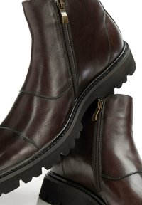 Botas de tobillo de cuero marrón oscuro con cremallera lateral, acabado texturizado, suela de goma con tacos y acentos lisos. Diseño clásico con detalles de costura.