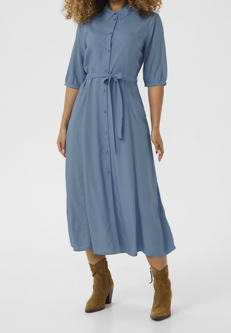 Femme portant une robe midi bleue à boutons avec une ceinture nouée et des manches jusqu'aux coudes, associée à des bottines marron en suède.