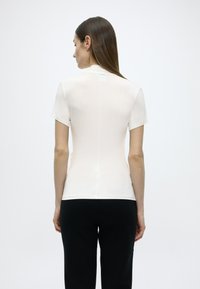 Κρεμ  πουκάμισο με κοντά μανίκια, εφαρμοστό σχέδιο, λεία υφή και μινιμαλιστικό στυλ. Στην πλάτη υπάρχει μια διακριτική ραφή.