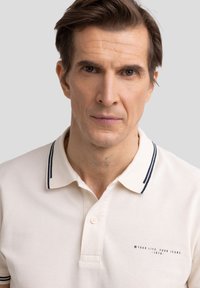 Polo majica v kremni barvi z teksturirano tkanino, z morsko modrim rebrastim ovratnikom in majhnim logotipom na levi strani. Dvoklapni gumb.