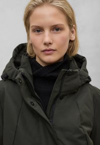 Olivově zelená bunda s kapucí na zip a patentní knoflíky, pod ní černý žebrovaný rolák. Hladká textura, minimalistický design.