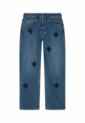 Jeans azules con patrones de flor de lis negra, llevados por una persona con zapatillas blancas y negras, de pie con las manos relajadas y en los bolsillos.