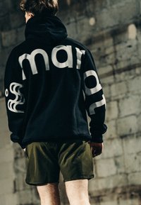 Persoon met een zwarte hoodie met grote witte letters en olijfgroene korte broek, staand voor een verweerde stenen muur.