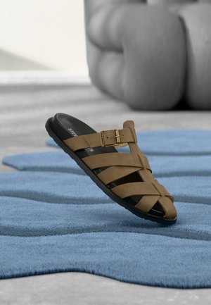 Bruine sandaal met riempjes en gesp op zwarte zool zwevend boven een gestructureerd blauw tapijt, met een vervaagde grijze sculptuur op de achtergrond.