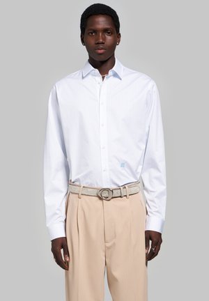 Homme portant une chemise blanche à manches longues boutonnée et un pantalon plissé beige avec une ceinture gris clair sur un fond gris uni.