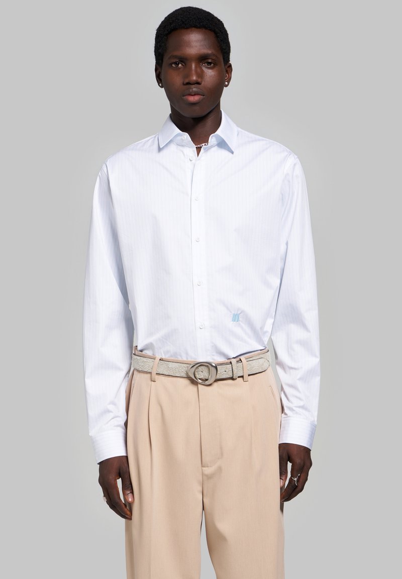Homme portant une chemise blanche à manches longues boutonnée et un pantalon plissé beige avec une ceinture gris clair sur un fond gris uni.