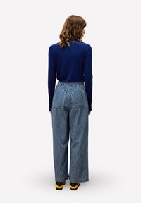 Person mit schulterlangem Haar, der ein navy-blaues Langarmoberteil, weiter gestreifte Hosen mit hinteren Taschen und Schuhe mit gelber Sohle trägt und von hinten zeigt.