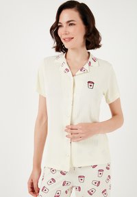 Ensemble de pyjama à manches courtes, de couleur jaune pâle, avec une texture côtelée. Le haut présente un col et des détails brodés en forme de tasse ; le pantalon arbore un motif de tasse assorti.