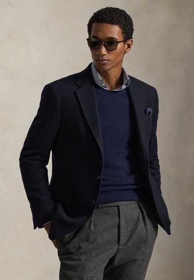 Man met donkere zonnebril, een marineblauwe blazer met pochet, een blauwe trui over een gestreept overhemd, en grijze broek tegen een effen achtergrond.