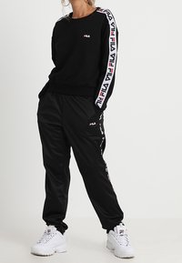 Sudadera negra con el logo de FILA en blanco y rojo a lo largo de las mangas, combinada con pantalones de chándal negros. Zapatillas blancas completan el conjunto.
