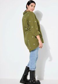 Olijfgroene oversized shirt met zilveren sterrenborduring, opgerolde mouwen, gecombineerd met lichtblauwe denim jeans en zwarte kniehoge laarzen.