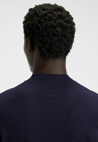Golf blu navy con colletto a coste, texture del tessuto liscia e un piccolo marchio sul retro. La vista posteriore mostra capelli corti e texturizzati.
