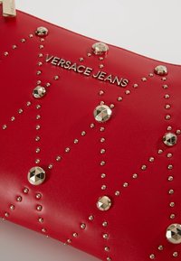 Sac en cuir rouge orné de clous dorés formant un motif, avec un logo métallique "VERSACE JEANS" au centre.
