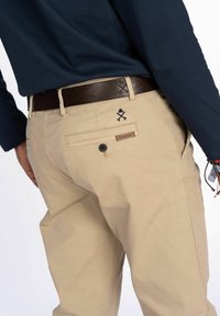 Mann in beigen Hosen mit Knopf an der Gesäßtasche, dunklem braunen Gürtel, navyblauem Langarmhemd und Handgelenkaccessoires vor weißem Hintergrund.