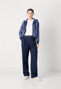 Giacca con cappuccio blu navy a motivo geometrico, abbinata a pantaloni larghi blu navy. Una camicia bianca e sneakers bianche completano l'outfit.