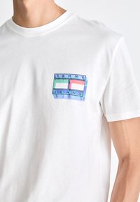 Camiseta blanca con un pequeño gráfico colorido en el pecho que muestra dos rectángulos y el texto "Tommy Jeans" en letras de bloque.