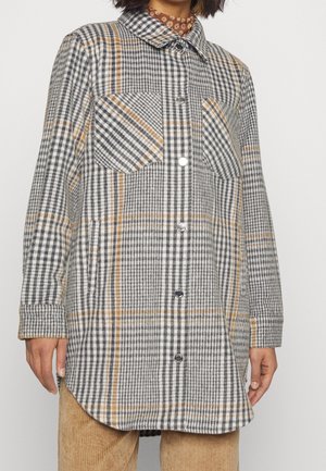 Femme portant un manteau à carreaux boutonné à manches longues gris, blanc et beige sur un pantalon en velours côtelé marron, sur un fond uni.