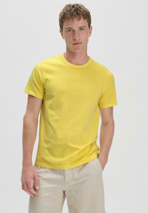 Gelbes Baumwoll-T-Shirt mit Rundhalsausschnitt und kurzen Ärmeln. Glatte Textur und lockere Passform. Kombiniert mit hellbeigen Hosen.