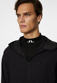 Svart zip-up jacka med huva, tillverkad av lätt material, har en glänsande svart logotyp på bröstet och en elegant, minimalistisk design.