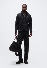 Άνδρας με μαύρη φόρμα Adidas και αθλητικά παπούτσια κρατάει μαύρη τσάντα γυμναστηρίου, στέκεται μπροστά σε απλό λευκό φόντο.