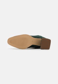 Zapato de ante verde con tacón cuadrado y suela de goma. La parte inferior presenta un patrón texturizado para mayor agarre, con un sutil acento marrón en el tacón.