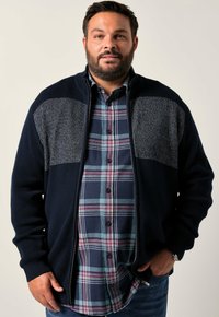 Marineblå zip-jacka med texturerad stickad design och grå paneler, lager-på-lager över en rutmönstrad skjorta i blå, grön och röd ton.