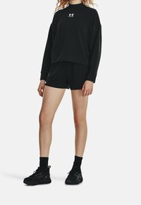 Schwarzes Sweatshirt und Shorts-Set aus Fleece-Material. Mit Rundhalsausschnitt und einem Logo auf der Brust. Kombiniert mit schwarzen Sportschuhen und Socken.