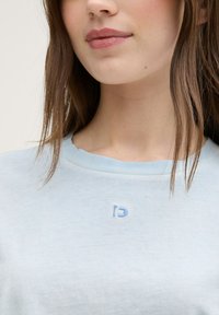 Cămiză din bumbac de culoare albastru deschis, cu un guler rotund. Prezintă un mic logo brodat în albastru deschis pe piept. Textură moale, croi lejer.