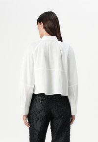 White collared shirt with long sleeves, featuring a slightly cropped back and fine stitching details, paired with black pants with floral texture.