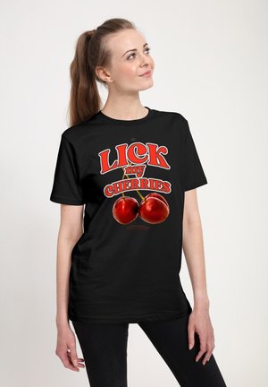 Žena s hnědým copem, oblečená v černém tričku s červeným nápisem "LICK my CHERRIES" a grafikou dvou červených třešní.