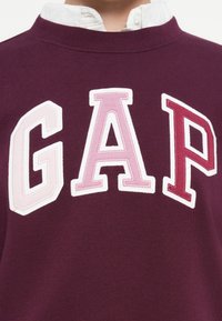 Bordo pulover z velikimi, šivanimi, univerzitetnimi črkami "GAP" v odtenkih roza in bele na prsih preko bele srajce s ovratnikom.