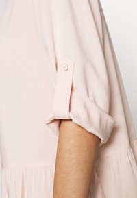 Blusa rosa chiaro con maniche arrotolate fissate con un bottone. Tessuto morbido e liscio, con una vestibilità ampia e fluida e orlo arricciato.