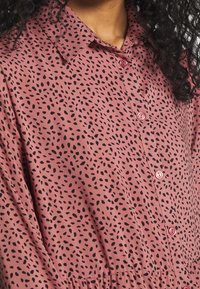 Rosa skjorta med knappar som har ett svart bladmönster, klassisk krage och textilstruktur. Knapparna har en matchande rosa nyans.