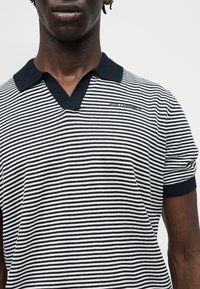 Άνδρας που φοράει κοντομάνικη πόλο μπλούζα με οριζόντιες ρίγες σε μαύρο και άσπρο, με μαύρο γιακά και μικρό λογότυπο στο στήθος.